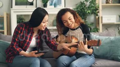 亚洲女人正在教她的非裔美国朋友在家里闲暇时间弹吉他。 一个女孩抱着